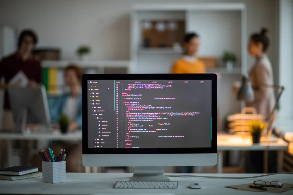 Close-up of a computer screen displaying colorful lines of code in an office. Three blurred people converse in the background, suggesting collaboration Mint Digital Solutions