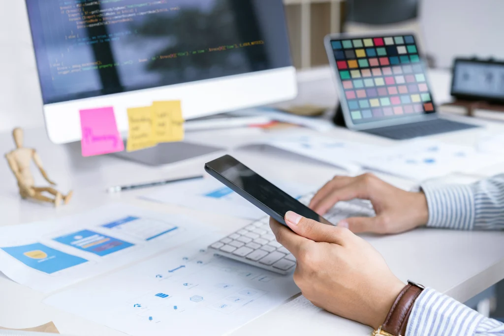 Person using a smartphone at a desk with a computer, colorful chart, and design sketches, conveying a tech-focused and creative work environment. Mint Digital Solutions