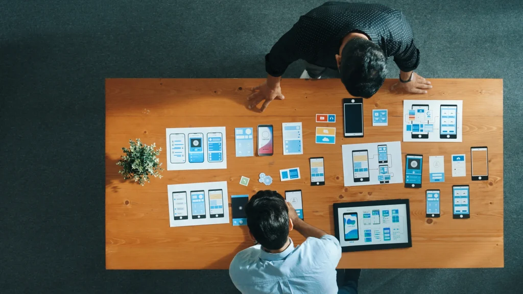 Two people collaborate on app design at a wooden table, viewing various smartphone layouts and prototypes. The setting is creative and focused. Mint Digital Solutions