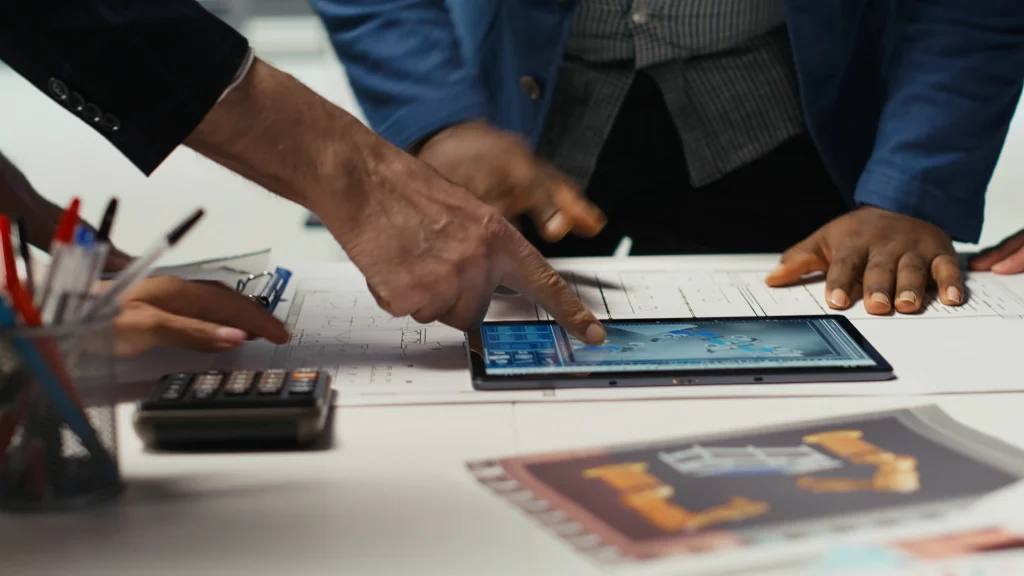 Hands of diverse professionals point to a tablet displaying blueprints on a cluttered desk with plans, a calculator, and office supplies, suggesting teamwork. mintdigitalsolutions