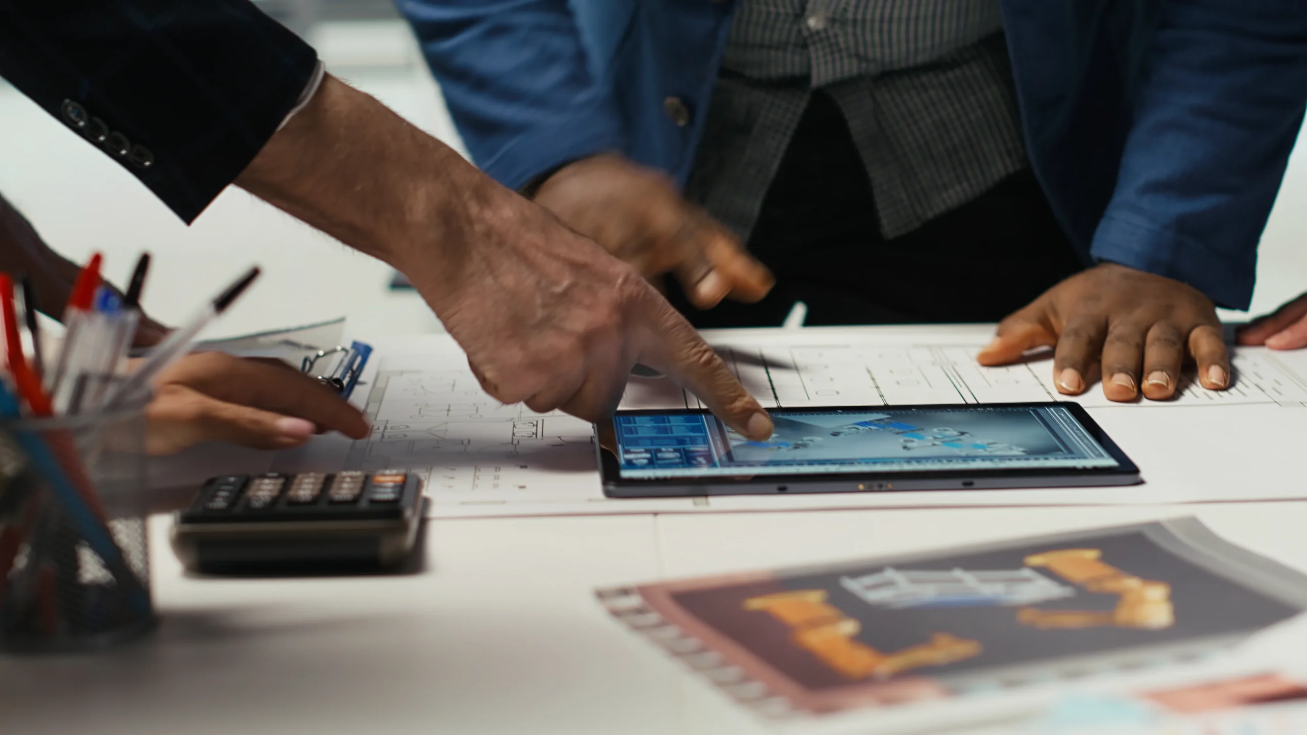 Hands of diverse professionals point to a tablet displaying blueprints on a cluttered desk with plans, a calculator, and office supplies, suggesting teamwork. mintdigitalsolutions