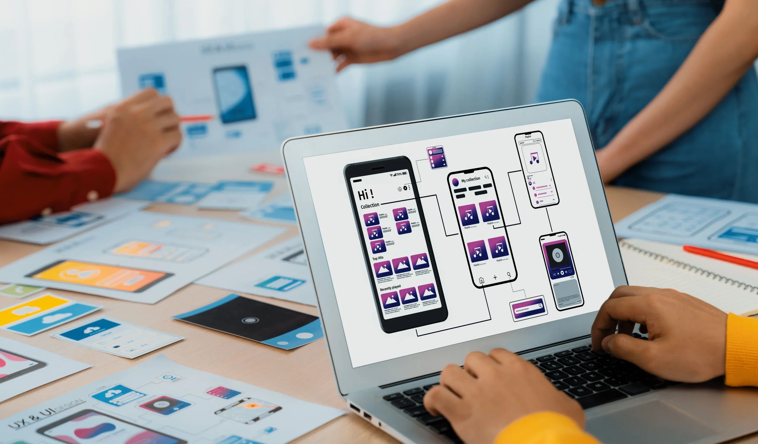 A person in a yellow sweater works on a laptop displaying a mobile app design. Two others discuss blueprints at a table filled with colorful design layouts. Mint Digital Solutions
