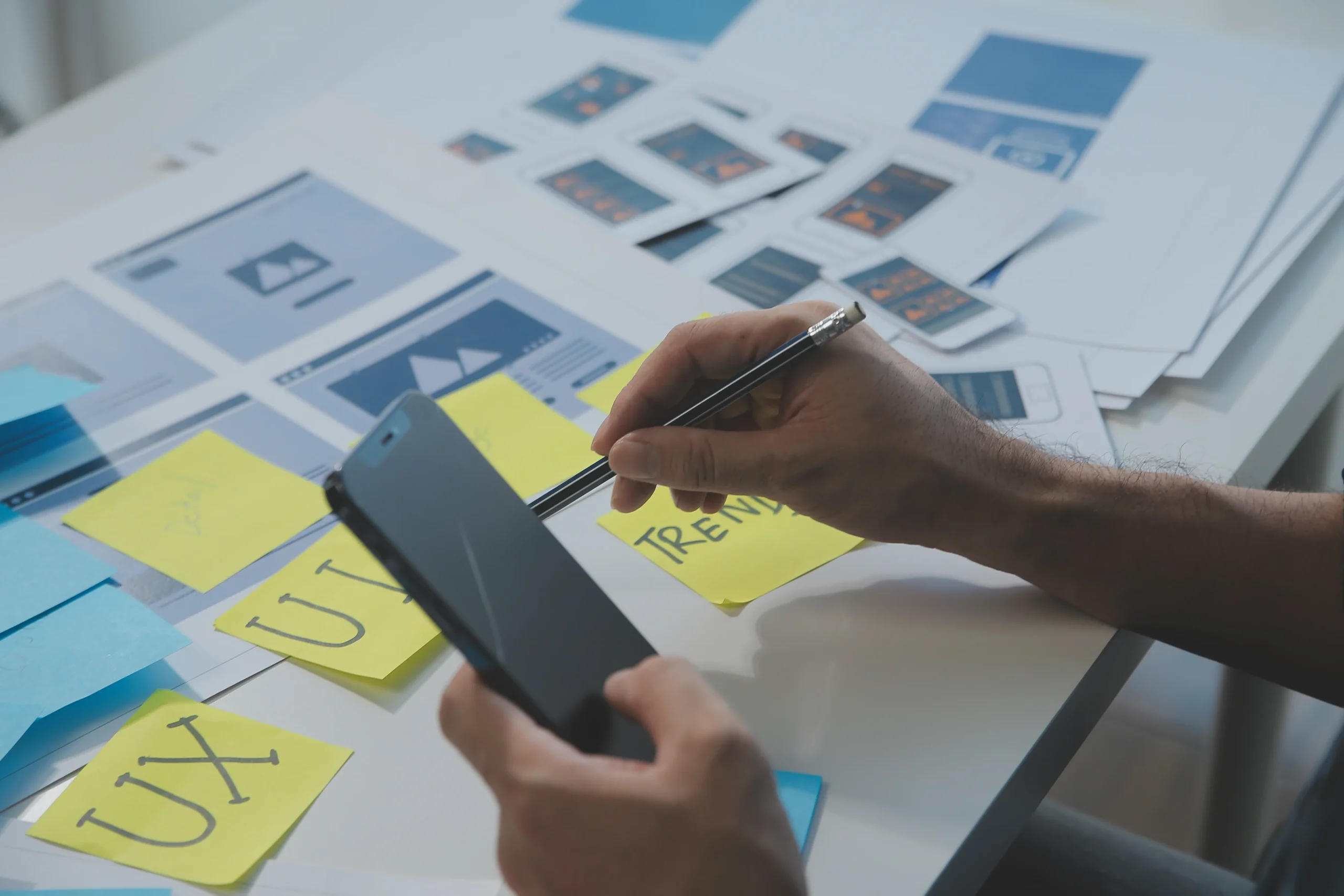 Hands working on UX design with wireframes and sticky notes labeled "UX" and "Trendy." A smartphone and pencil are used, indicating focus and creativity. mint digital solutions