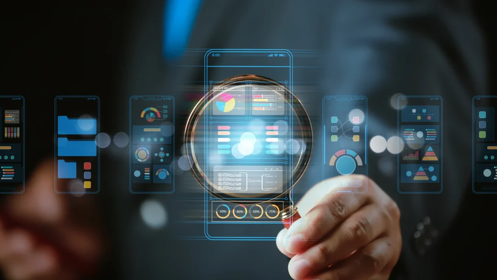 A hand holds a magnifying glass over digital smartphone screens displaying colorful graphs and data, symbolizing data analysis and technology focus. mint digital solutions