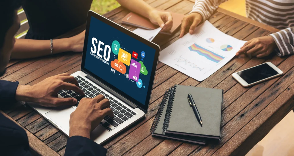 Three people work at a wooden table. One types on a laptop displaying "SEO" and colorful icons, while others discuss graphs and charts. A smartphone and notebook are nearby. mintdigitalsolutions
