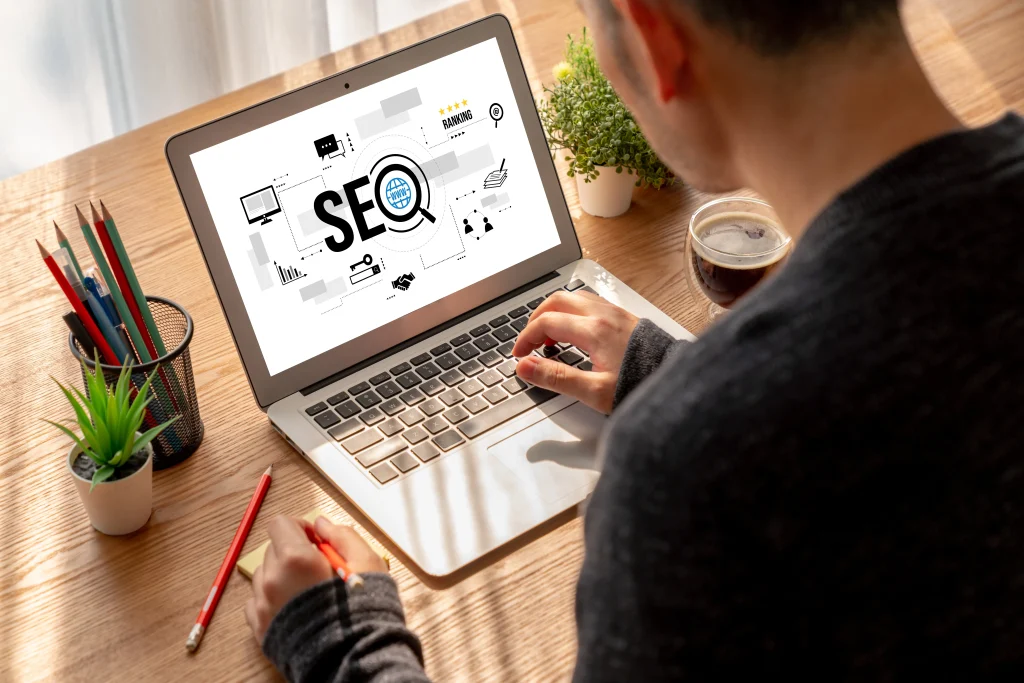 A person working on a laptop displaying SEO graphics, surrounded by a plant, colored pencils, and a cup of coffee on a sunlit wooden desk. mintdigitalsolutions