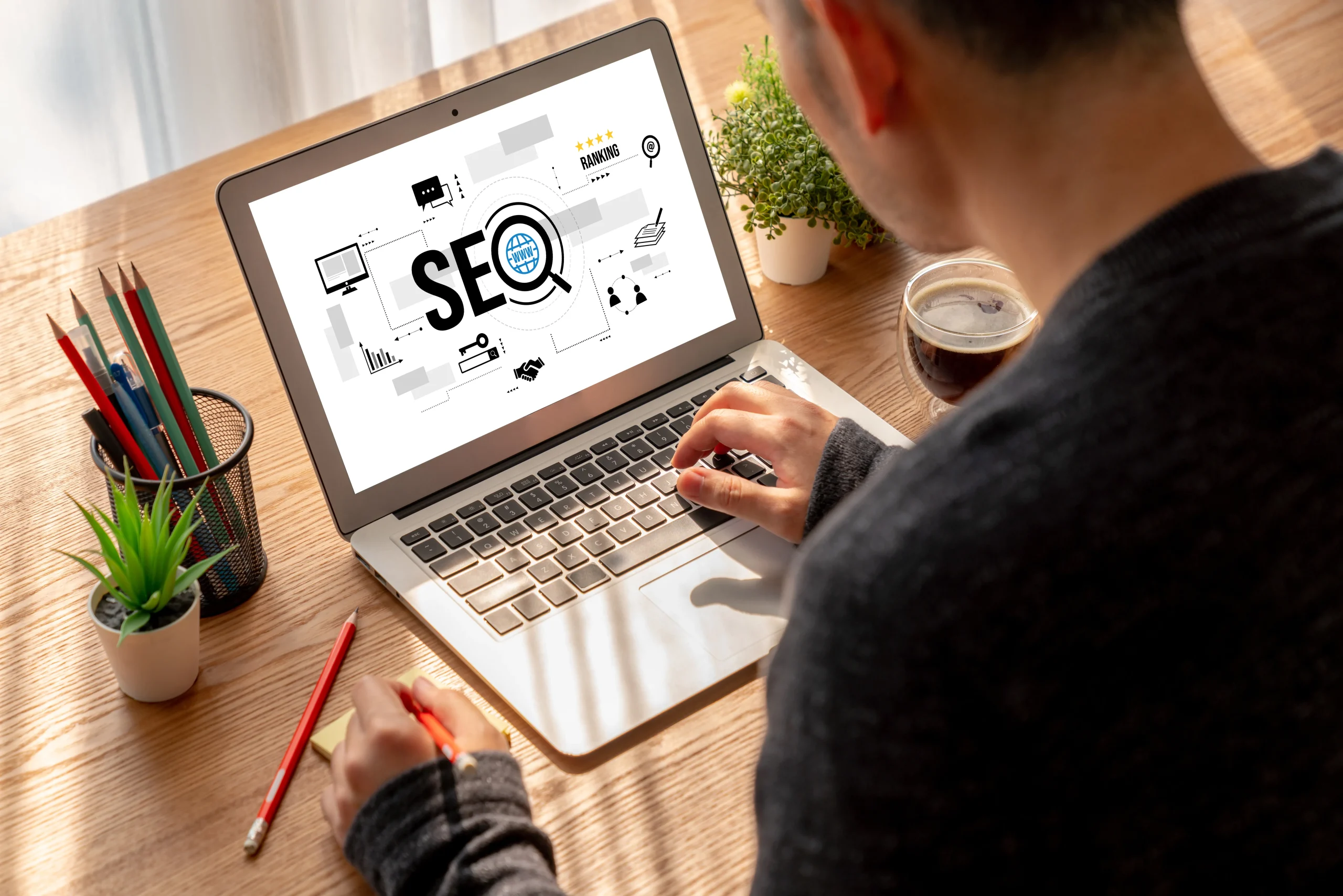 A person working on a laptop displaying SEO graphics, surrounded by a plant, colored pencils, and a cup of coffee on a sunlit wooden desk. mintdigitalsolutions