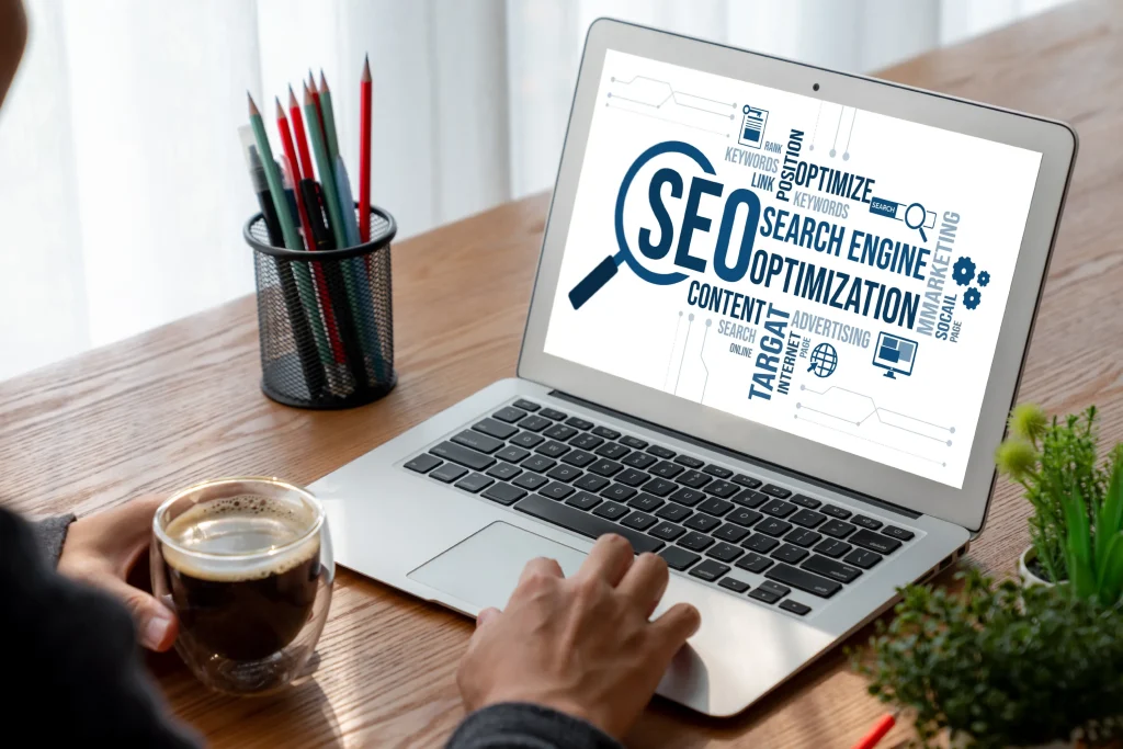 A person types on a laptop displaying a word cloud focused on "SEO" and related terms. Nearby, a hand holds a coffee cup. The setting is casual and focused. mintdigitalsolutions